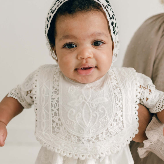 A baby wearing a white crochet bonnet and an Adeline Bib smiles while being supported by an adult. The baby, dressed in a Christening Dress, has dark eyes and appears cheerful, with one hand visible holding onto the adult.