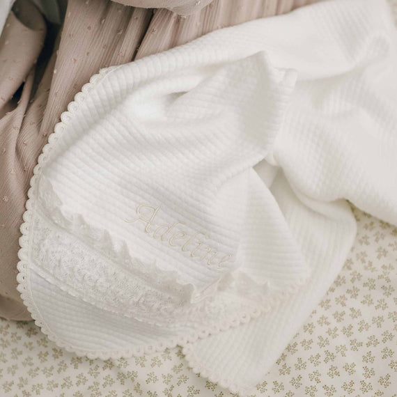 The Adeline Personalized Blanket, a soft, white textured handmade blanket with scalloped edges, lies folded on a delicate beige-patterned fabric. It boasts intricate lace detail and is embroidered with a name in cursive lettering, creating a cherished keepsake. Nearby rests a taupe-colored fabric adorned with small raised dots.