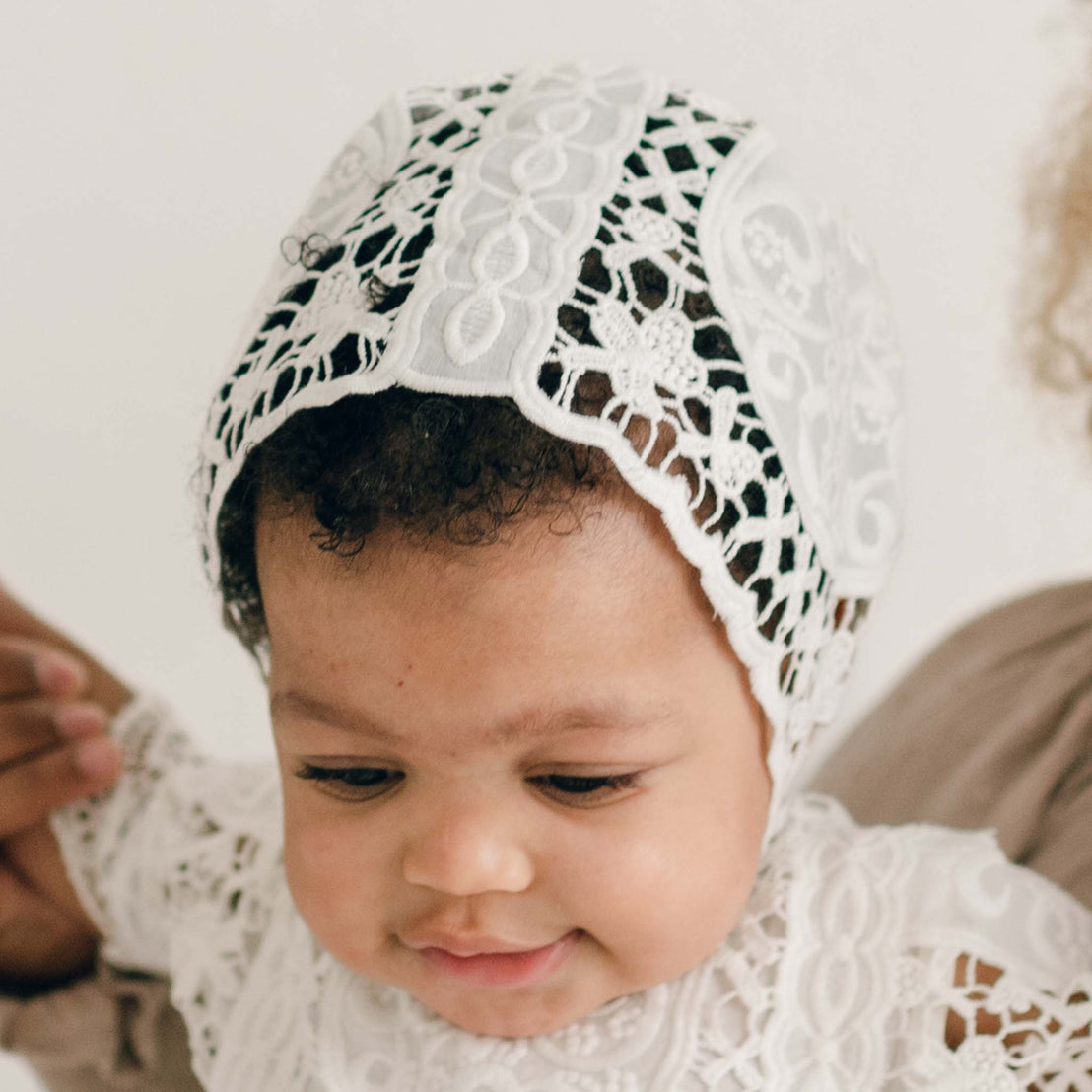 A baby wearing the Adeline Lace Bonnet with a silk ribbon and matching outfit gazes down thoughtfully. An adult's hand tenderly rests on the baby's side, offering support and care. The neutral background accentuates this touching moment.