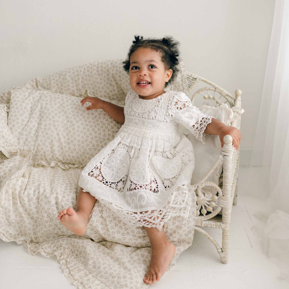 A young child with curly hair sits on a cushioned wicker bench, wearing the Adeline Lace Dress & Bloomers made of 100% cotton lace. The child smiles and holds onto the bench edge, one foot slightly lifted. The background is soft and neutral with light curtains and a floral-patterned blanket.