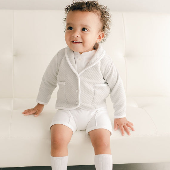 Baby boy sitting on a white couch and wearing the grey Asher 3-Piece Suit, including a folded collar jacket, white pants and a onesie with buttons.