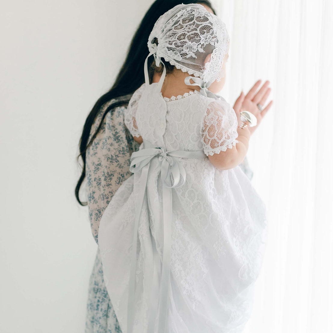 A young child in a delicate white Olivia Lace Bonnet and dress stands facing a bright window, the light softening the details of the lace and fabric, ready for her baptism.