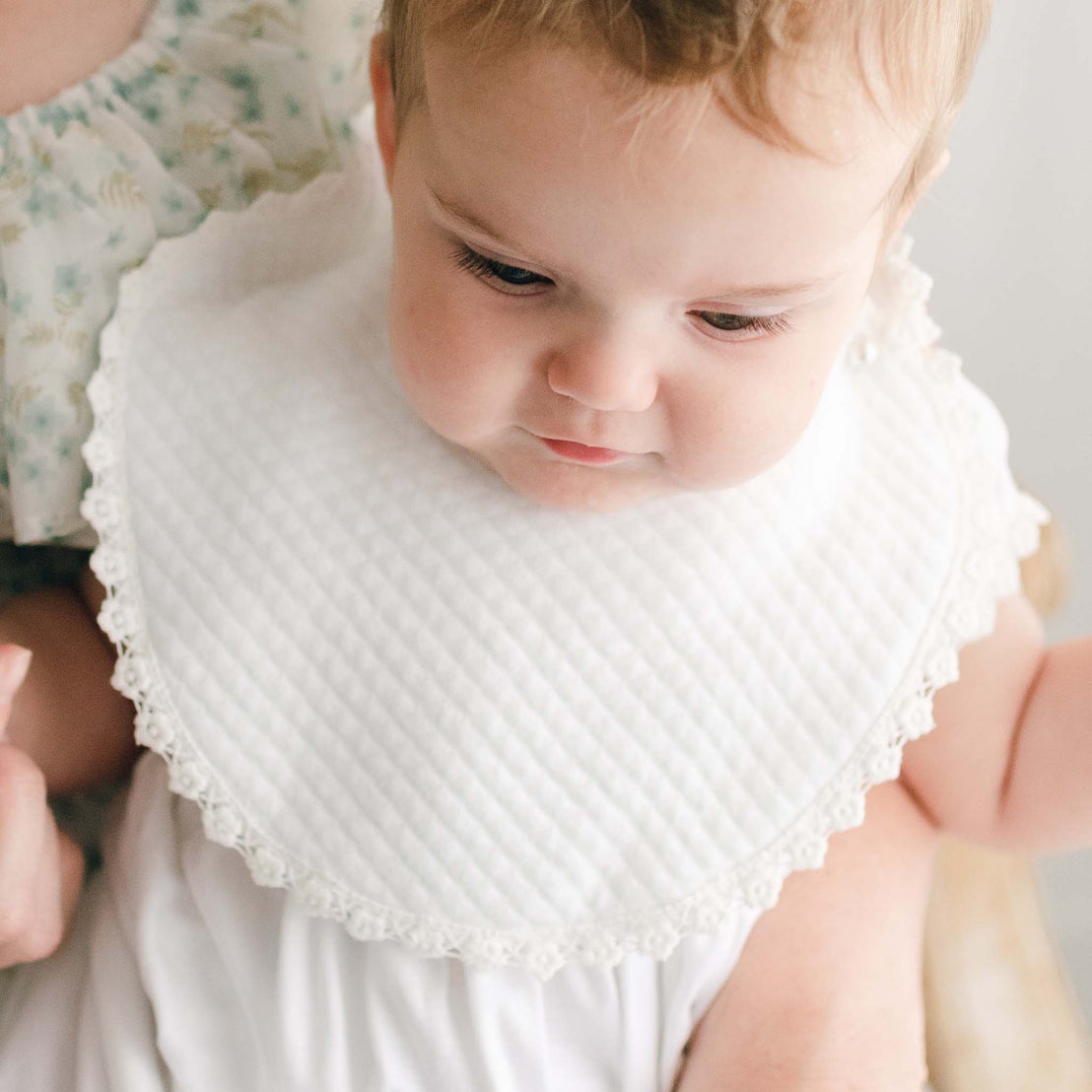 A newborn baby girl looks down away from the camera, wearing the Ella christening bib.