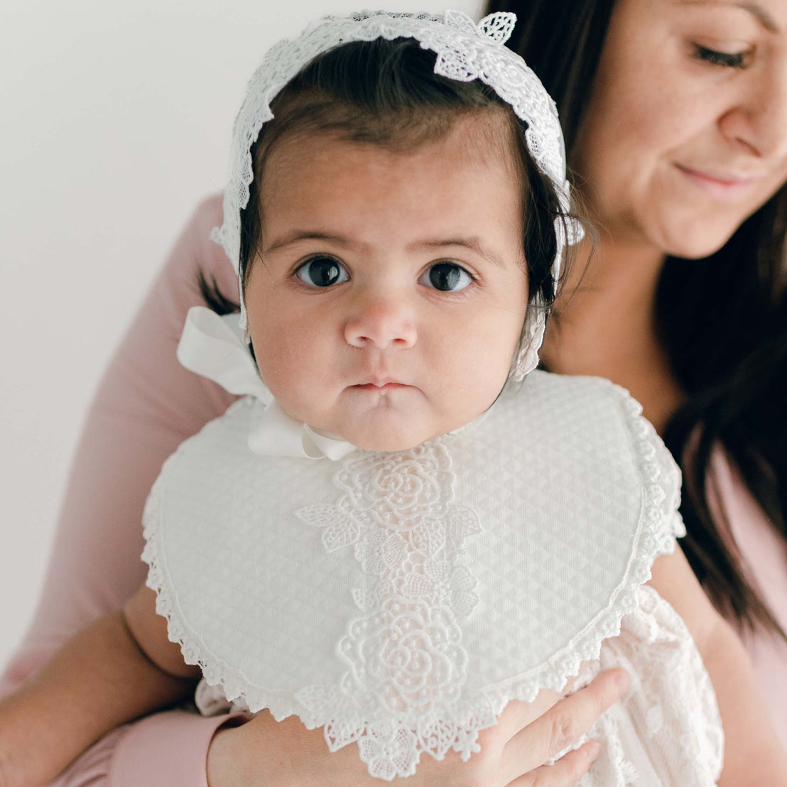 A baby with big expressive eyes, dressed in a bonnet, is held securely by a woman wearing a pink blouse.