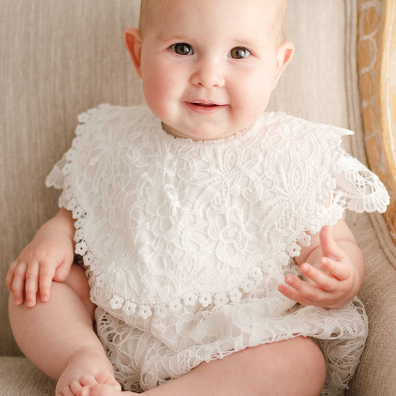 A newborn baby girl wearing the Lola Lace Bib over the Lola Bubble Romper, looking at the camera with a slight smile. The bib features an intricate lace overlay and cotton lace accents along the edge.