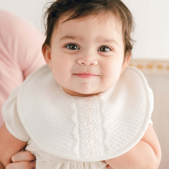 A close-up of a joyful baby with big eyes and an Emma Bib, being held by an adult in a pink shirt.