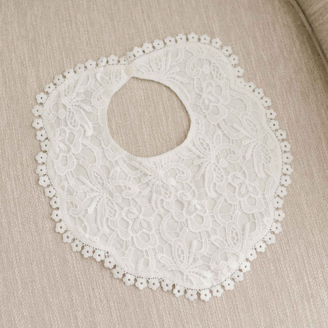 Top-down view of the Lola Lace Bib on linen fabric, featuring a delicate lace overlay, cotton lace trim, and a button closure at the back.