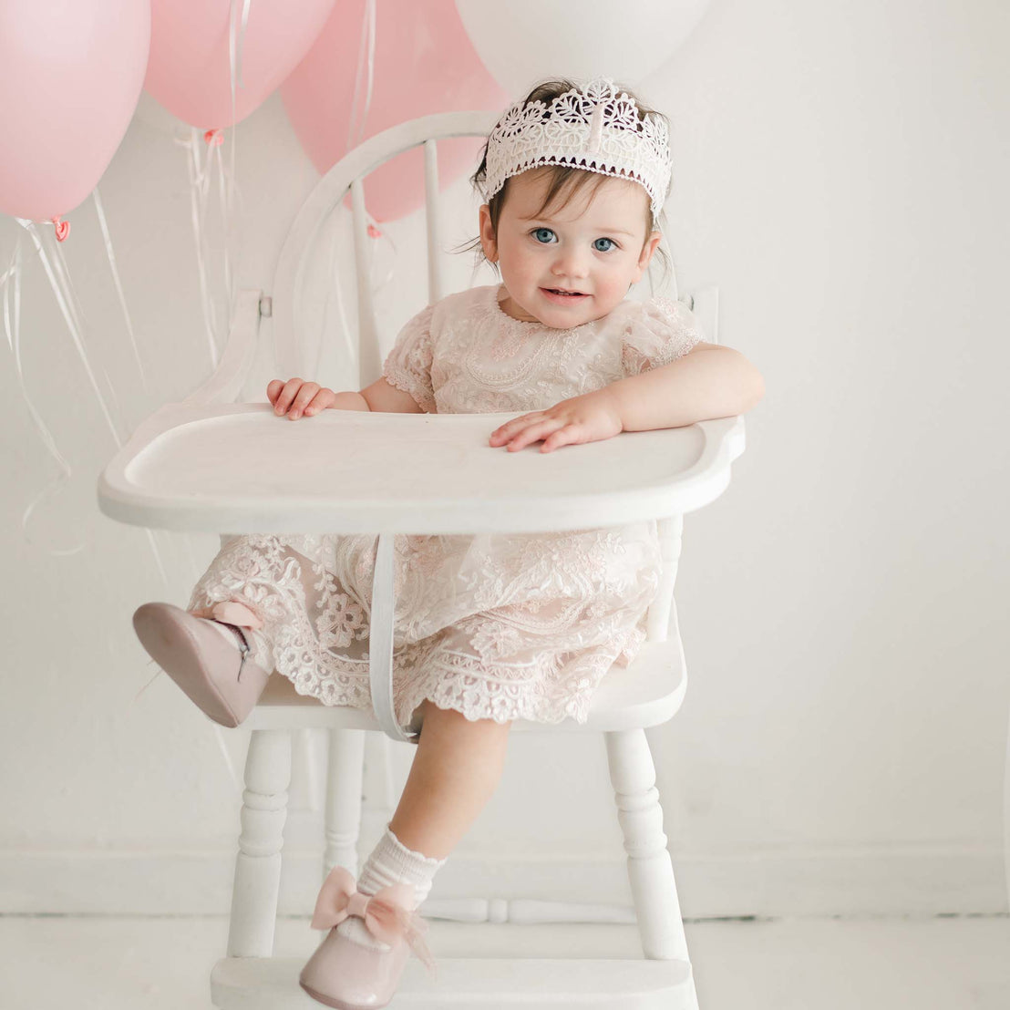 A toddler wearing a lace dress and the First Birthday Crown sits in a white high chair. Pink and white balloons are tied to the chair. She looks at the camera with a smile, her hand resting on the tray in front of her, and she is wearing light pink shoes with white socks.