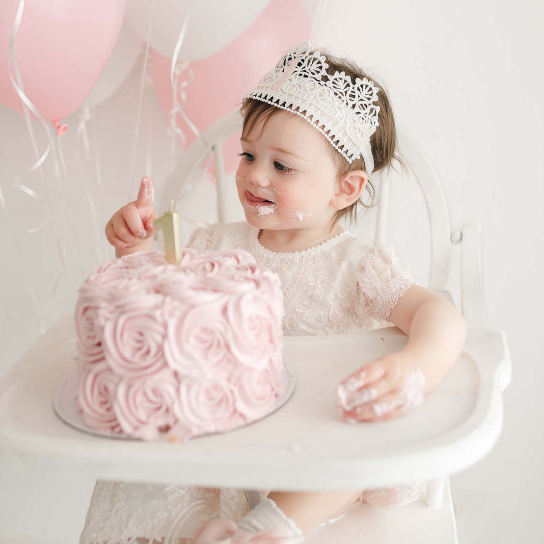 A toddler, joyfully seated in a high chair, is adorned with a First Birthday Crown and a white dress while eagerly touching a large pink frosted cake decorated with rosettes. In the background, pink and white balloons can be seen. The child has some frosting on their face and hands.