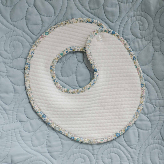 Flat lay of the Petite Fleur Bib made with soft ivory quilted cotton and trimmed with floral cotton in blue and yellow, featuring a button closure at the shoulder.