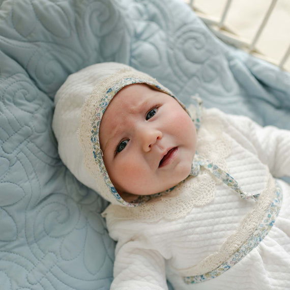 Baby wearing the Petite Fleur Quilted Bonnet, made from soft quilted cotton and trimmed with floral cotton and ecru lace, resting on a light blue quilted blanket.