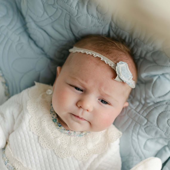 Baby wearing the Petite Fleur Linen Flower Headband, featuring a soft lace band with a light blue linen flower, resting on a quilted blue blanket.