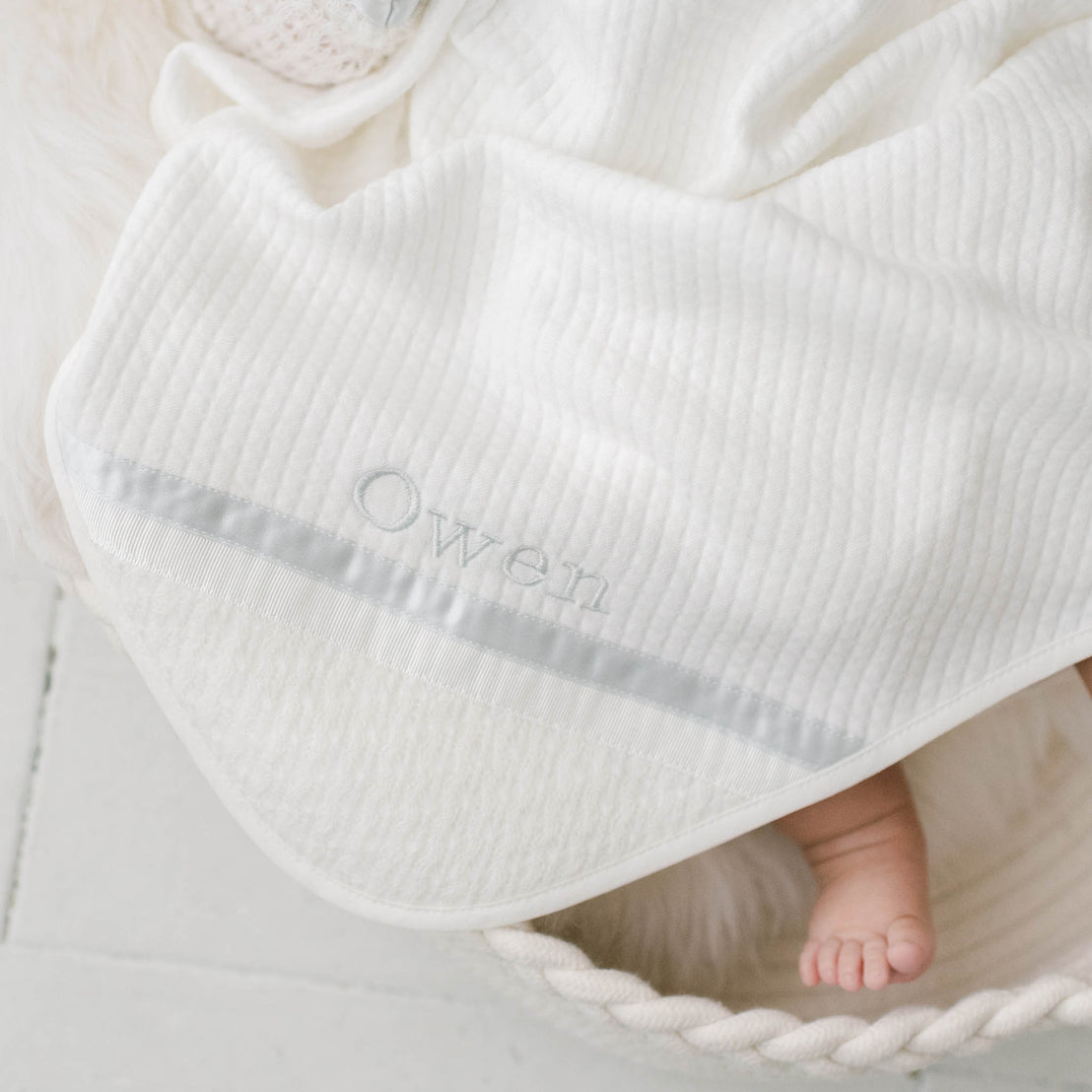 Photo of the corner of the Owen Personalized Blanket. The corner features an ivory knit and the name "Owen" embroidered with light blue thread