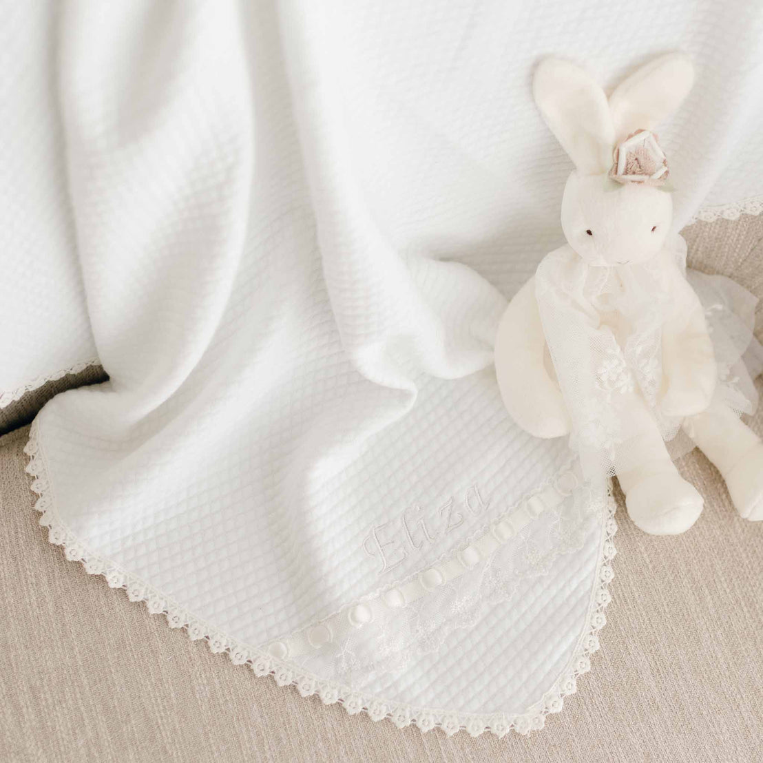 An upscale, Eliza Silly Bunny Buddy | Pacifier Holder with a lace bow sits atop a soft, textured white christening blanket with delicate lace trim on a light beige background.