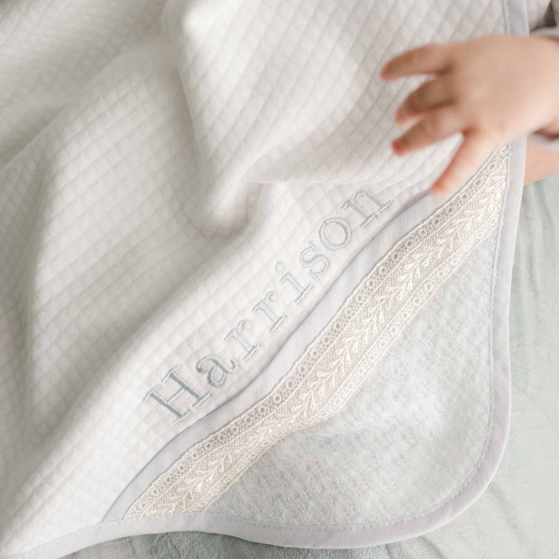 Photo of a baby hand touching a Harrison Personalized Blanket made with a plush quilted white cotton and trimmed with light blue linen. The accent corner features ivory Venice lace. On the corner of the blanket is the name "Harrison" embroidered in light blue thread