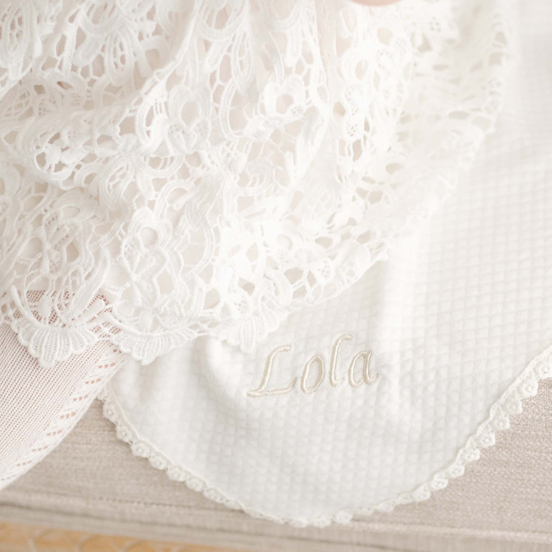 Close-up of the Lola Personalized Blanket in light ivory cotton with lace trim, featuring 'Lola' embroidered on the corner, paired with a delicate lace christening gown.