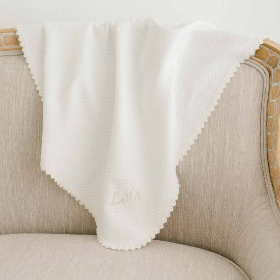 Lola Personalized Blanket draped over a beige chair, made from soft light ivory cotton with delicate lace trim and 'Lola' embroidered on the corner.