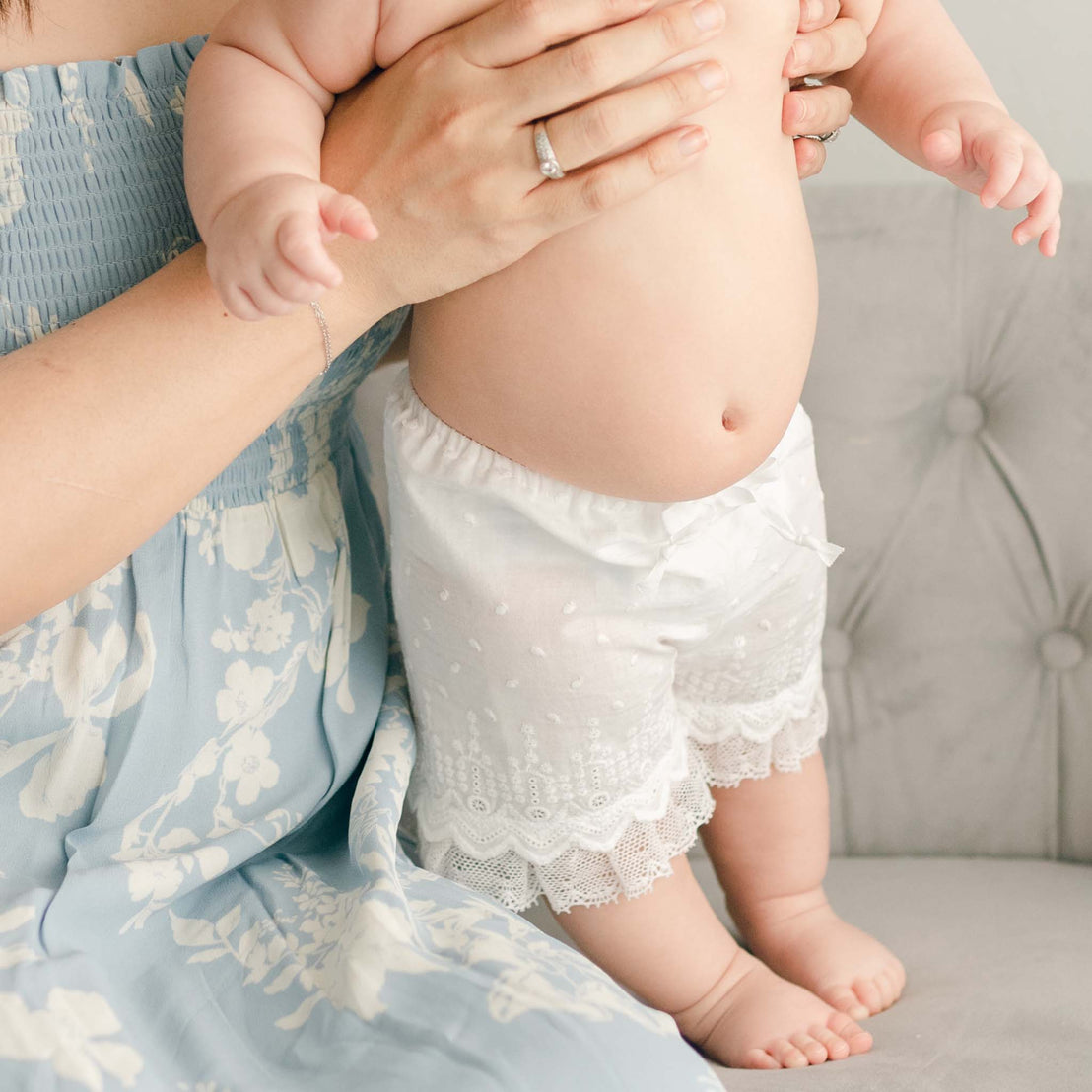 A mother in the **Emily Dress & Bloomers | Red Sash** cradles her baby at the christening; the focus is on the baby, dressed in white shorts, and her embrace.