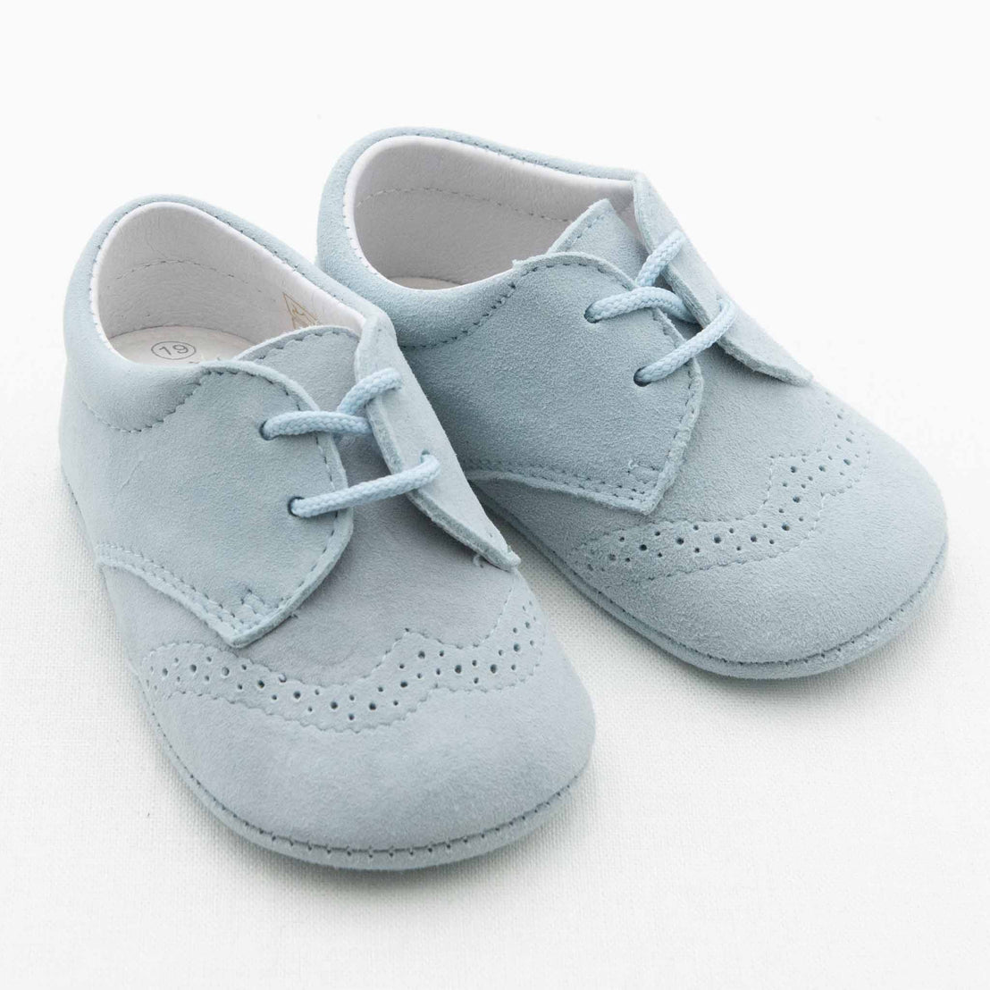 A pair of Sky Blue Suede Shoes, small and light blue with decorative perforations. Handmade in Spain, these baby shoes feature laces and a soft, comfortable design perfect for infants. They are placed side by side against a white background.