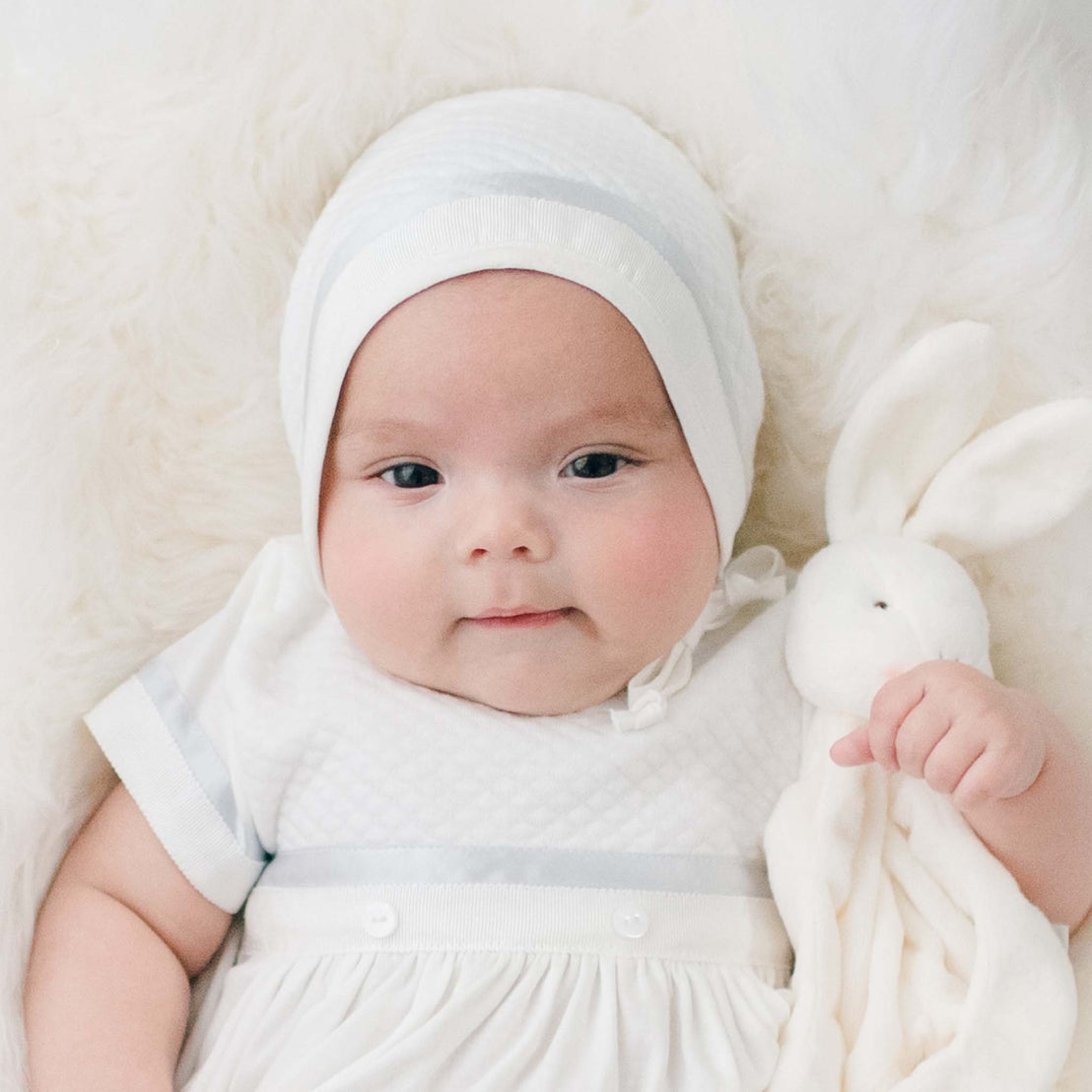 Newborn baby boy sitting in a white fur-lined crib. He is wearing the Owen Layette and Quilted Newborn Bonnet. He is also holding the Owen Silly Bunny Buddy