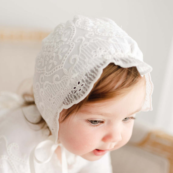 A toddler with light brown hair wears an Eliza Bonnet suitable for a christening and gazes downward with a gentle expression, set against a soft, neutral background.