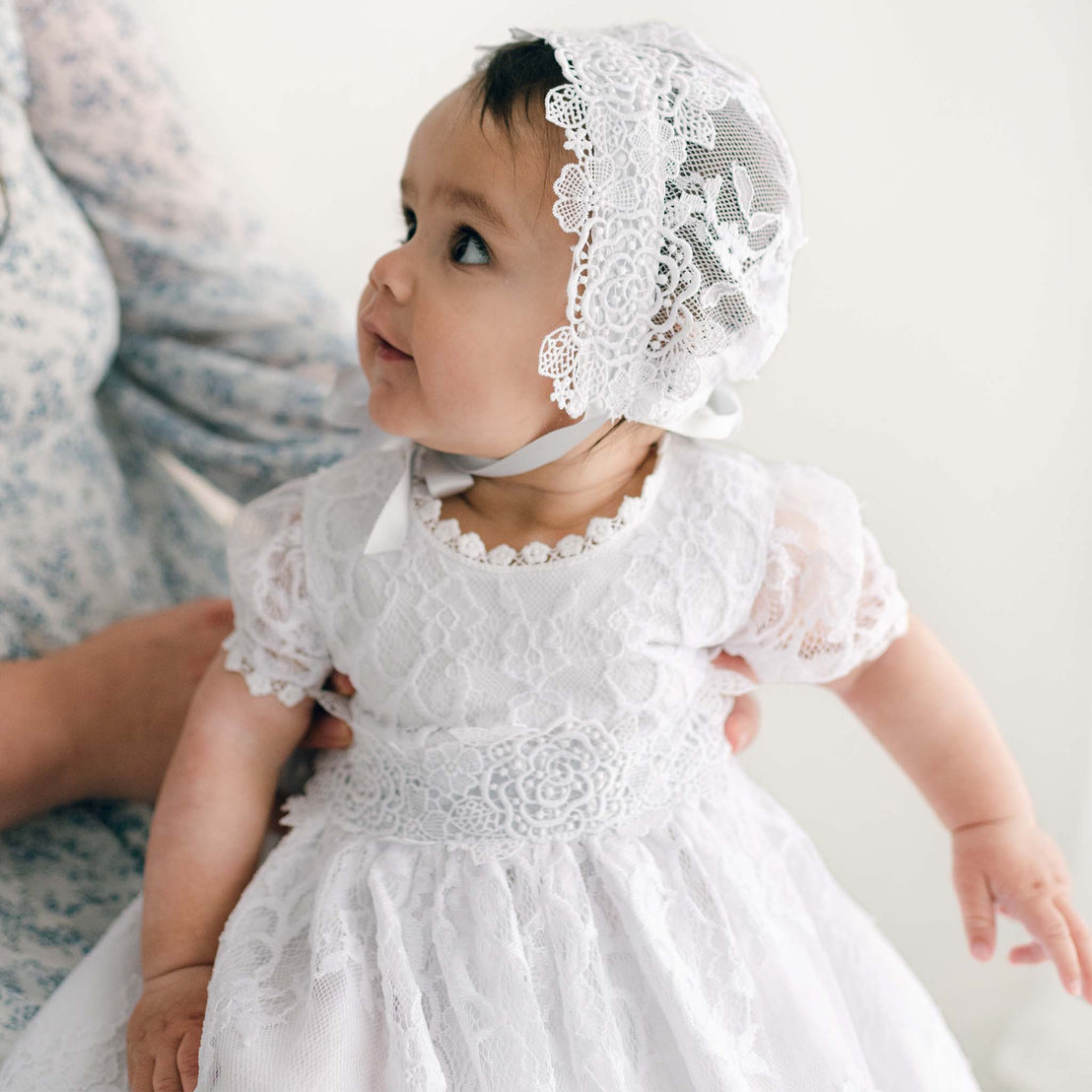 A baby girl in a white Olivia Lace Bonnet and dress looks up curiously, held securely in the arms of an adult wearing a blue shirt, ideal for a christening or as a boutique baby gift.