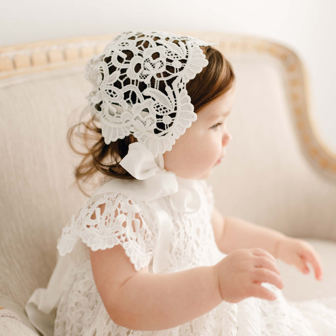 Baby girl wearing the Lola Lace Bonnet with intricate white lace and satin ribbon ties, seated in a cream-colored chair.