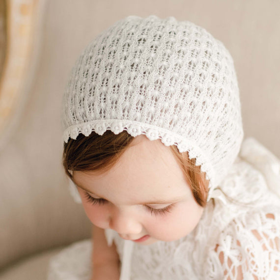 Close-up of a baby girl wearing the Lola Knit Bonnet, made from soft light ivory knit acrylic with delicate cotton lace trim and silk ribbon ties.