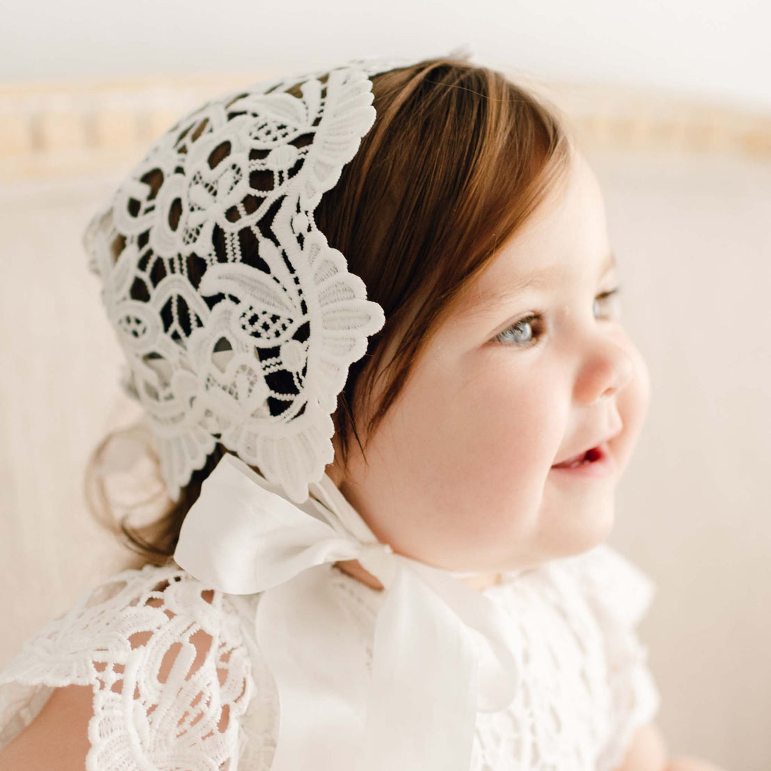 Close up of a baby girl wearing the Lola Lace Bonnet designed with embroidered light ivory lace and cotton floral edge lace at the back. The baby girl is smiling slightly as she looks to the side.