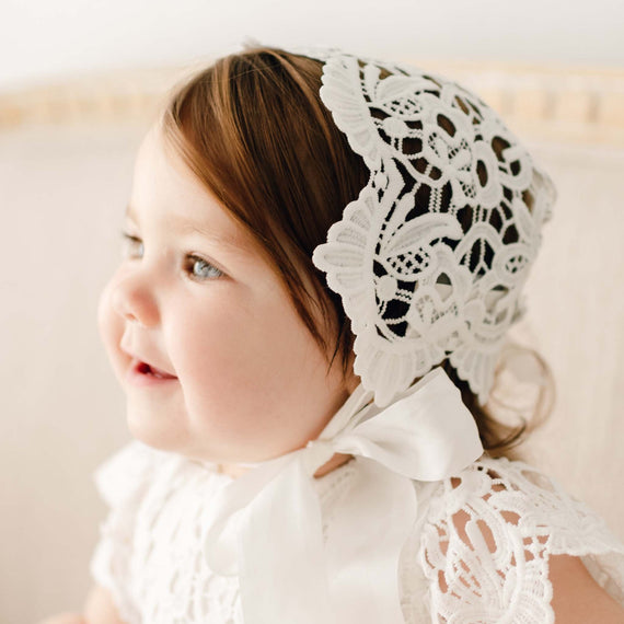 Baby girl with brown hair smiles gently, wearing the Lola Lace Bonnet and matching lace dress, in soft natural light that enhances the delicate lace details.