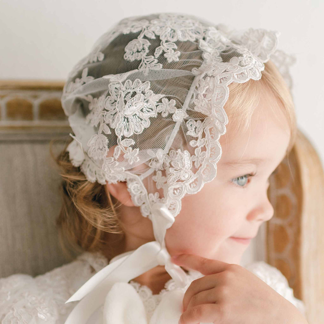 Close up detail of baby girl in profile wearing the Penelope lace christening bonnet. She is sitting in a beige upholstered chair. The background is softly blurred.