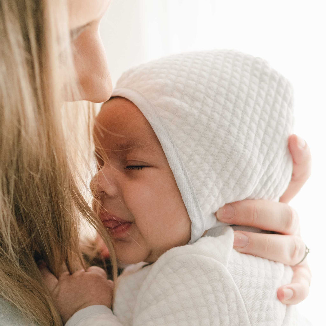 A close up detail of a mother with her newborn baby boy wearing a Harrison Quilted Bonnet made with soft quilted cotton in white and trimmed in a light blue linen