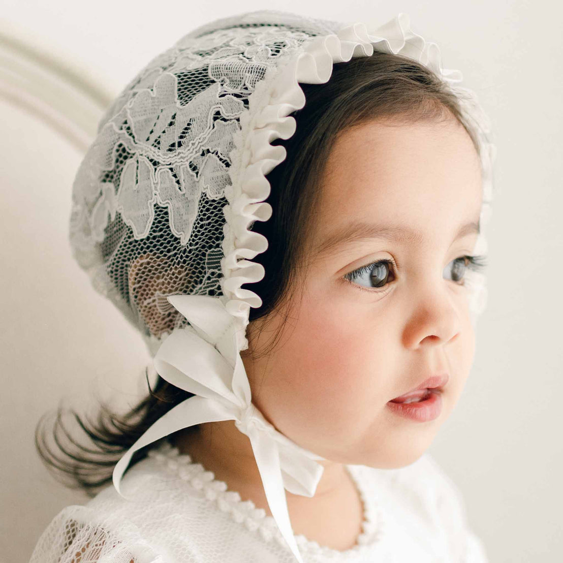 A toddler looks aside away from the camera. She is wearing the Victoria Lace Bonnet. The background is softly blurred and accentuates the details of the lace bonnet.