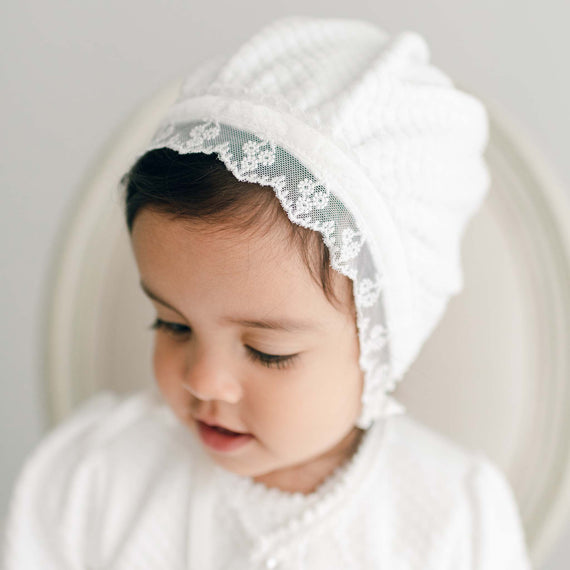 Baby girl wearing the Emily Quilted Cotton Bonnet made from a soft quilted cotton in a shade of white. The bonnet is also adorned with light ivory floral lace trim and white velvet ribbon.