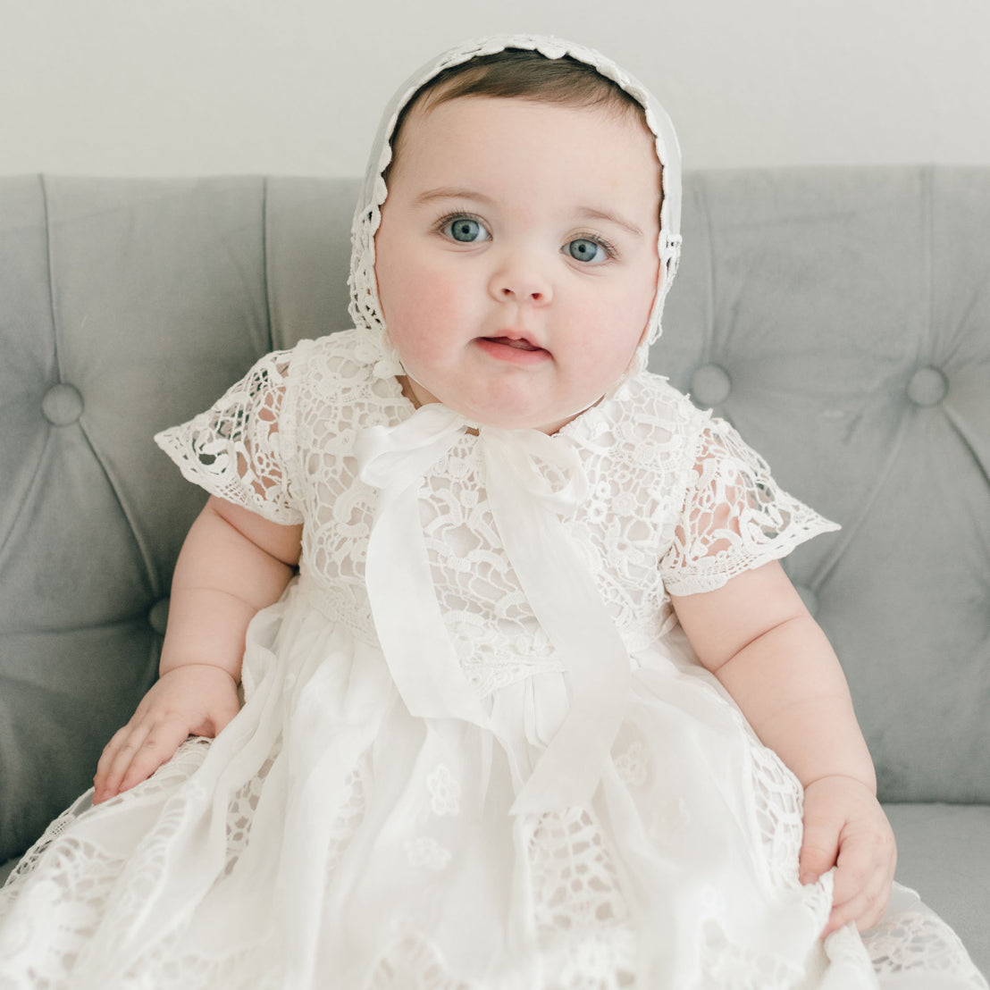 A baby wearing an ivory lace dress and the Grace Lace Bonnet sits on a gray tufted sofa. The little one has bright blue eyes and a neutral expression, with hands resting gently on the soft fabric.