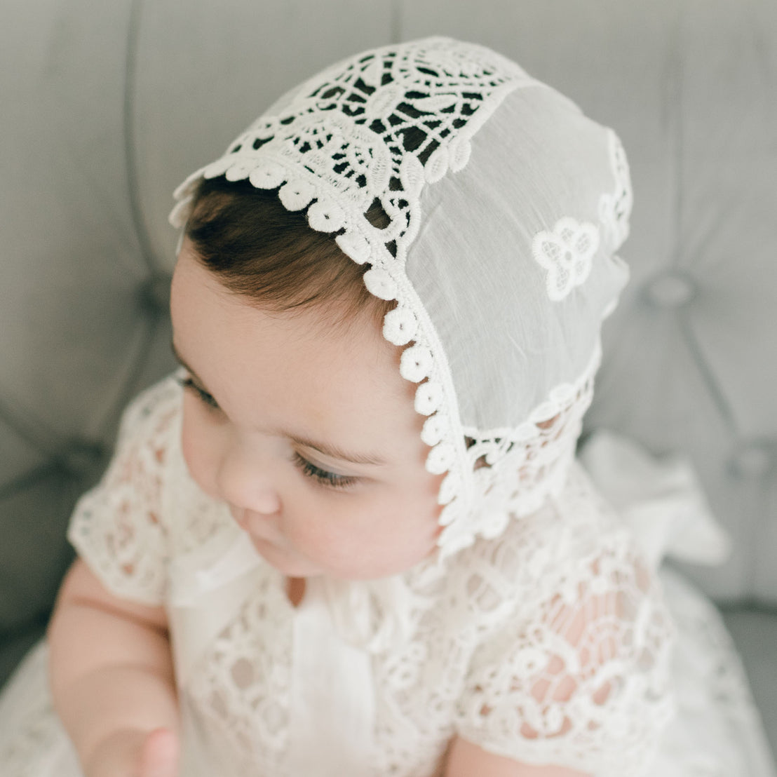 A baby, adorned in a Grace Lace Bonnet and a white lace dress, sits on a gray tufted surface, gazing downward. The intricate ivory lace design of the bonnet contrasts with the soft fabric of the dress, creating a classic, elegant look.