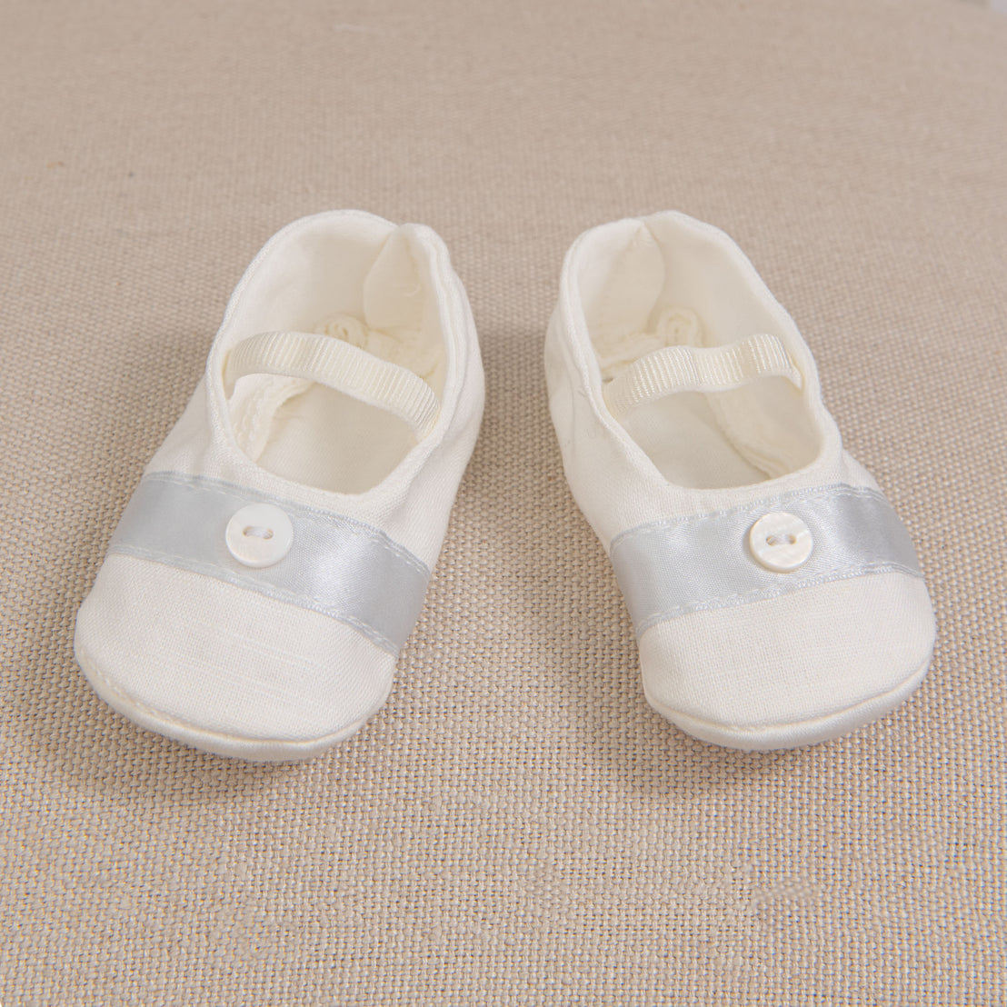 A pair of Owen Linen Booties displayed on a beige woven fabric surface. The booties are made from a linen/cotton blend in light ivory with a blue silk ribbon trim and button detail.