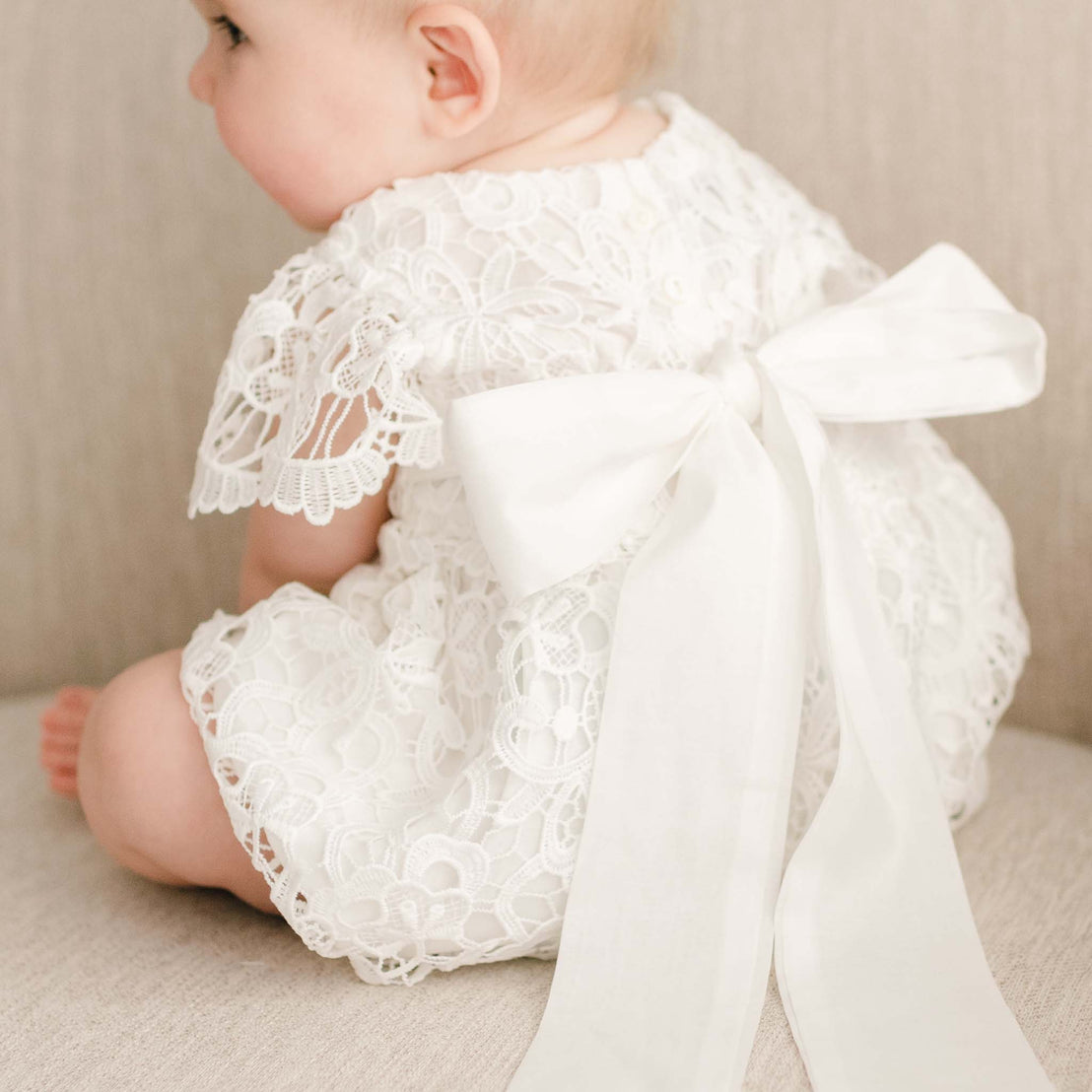 Back view of a baby girl sitting on a linen chair, wearing the Lola Lace Bubble Romper, with a large sash tie fastened into a bow.