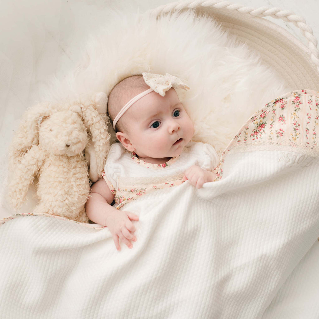 Newborn baby wearing the matching Eloise Layette Gown and Eloise Headband underneath the Eloise Personalized Blanket in the "Blush color"'.