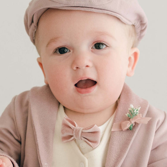 Closer detail of baby boy standing his mother's lap. He is wearing the Ezra 4-Piece Mauve Suit with matching Newsboy Cap, mauve velvet bow tie and boutonniere