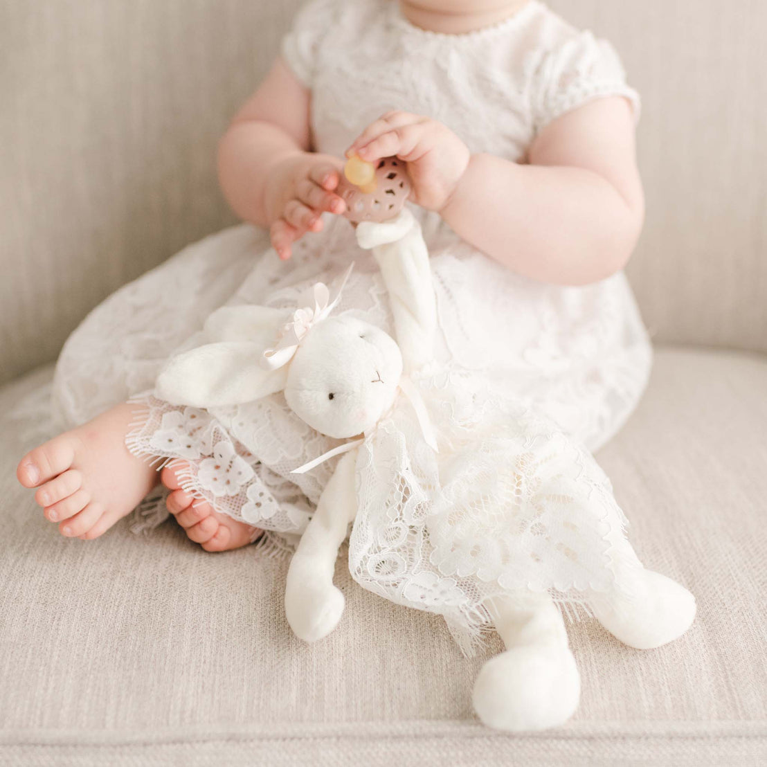 A baby holding the Victoria Silly Bunny Buddy and sitting on a beige sofa. She is dressed in the Victoria Puff Sleeve Christening Dress.