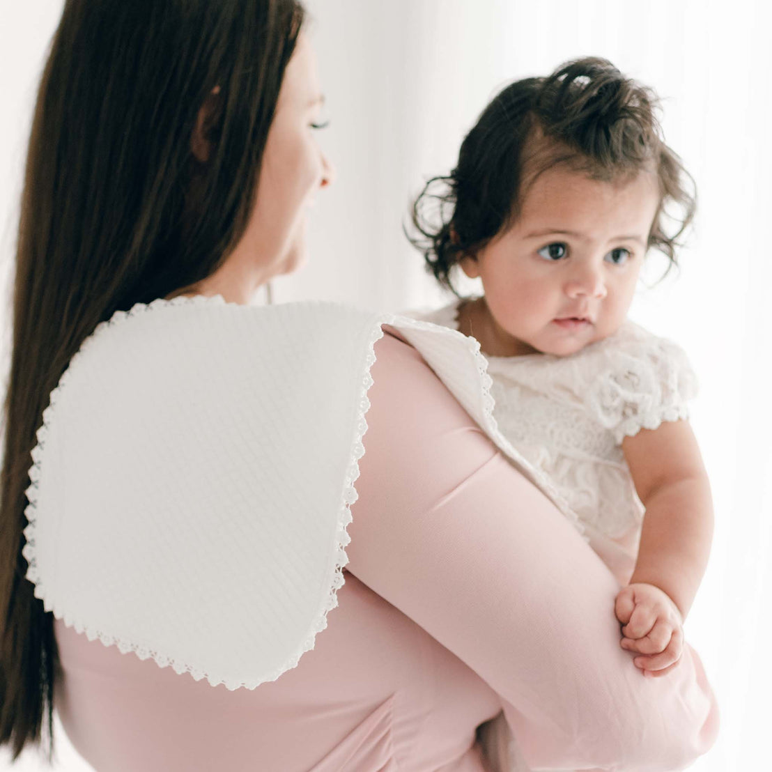 A woman in a pink boutique dress holding a baby girl with curly hair. The baby is dressed in a Ivory Christening Burp Cloth and they stand against a bright, softly lit backdrop.