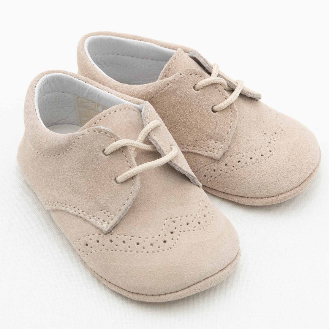 A pair of Camel Suede Shoes with decorative perforations, white interior lining, and white laces. These handmade suede shoes have a classic, formal design suitable for infants. The laces are tied in simple bows, and the Baby Beau & Belle boys shoes are displayed on a white background.