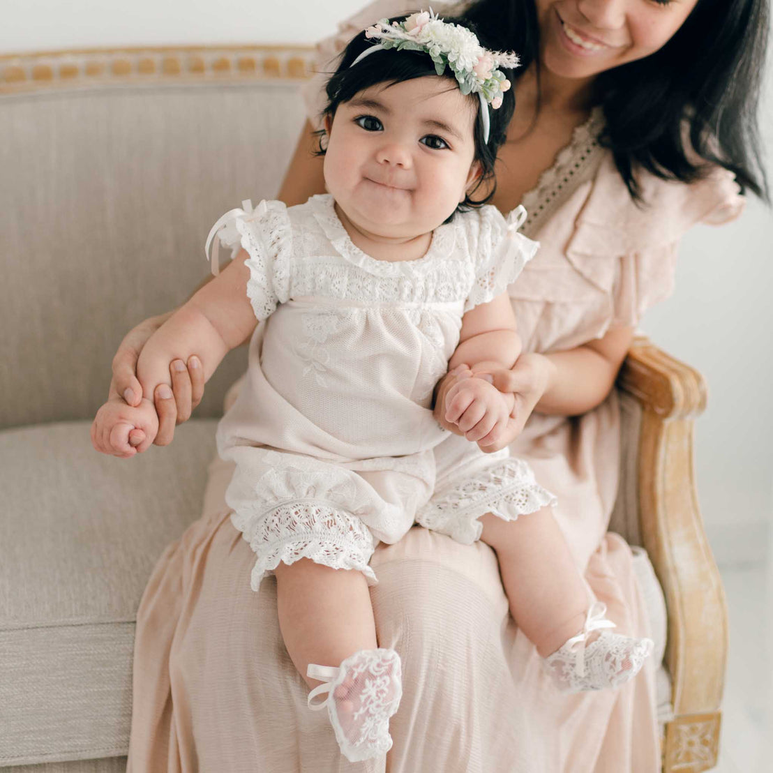 A baby girl dressed in the Charlotte Romper with matching booties and a floral headband sits on her mother's lap. The mother, partially visible, is wearing a light-colored dress and is holding the baby's hands while they both sit on a beige upholstered chair.