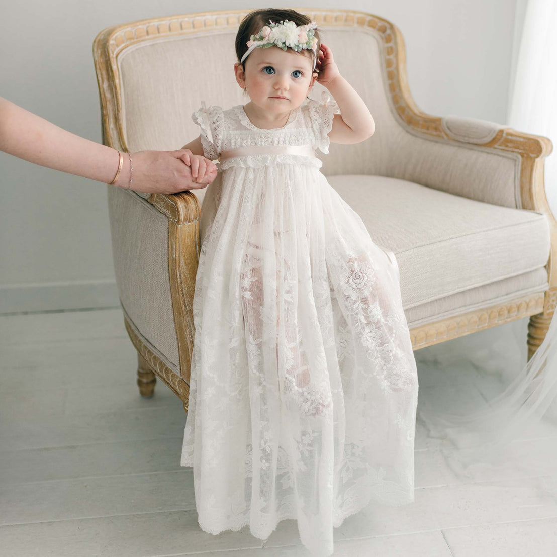 Baby girl wearing the Charlotte Flower Headband and lace gown, sitting on an upholstered linen chair while holding her mother's hand, her other hand gently touching her head.