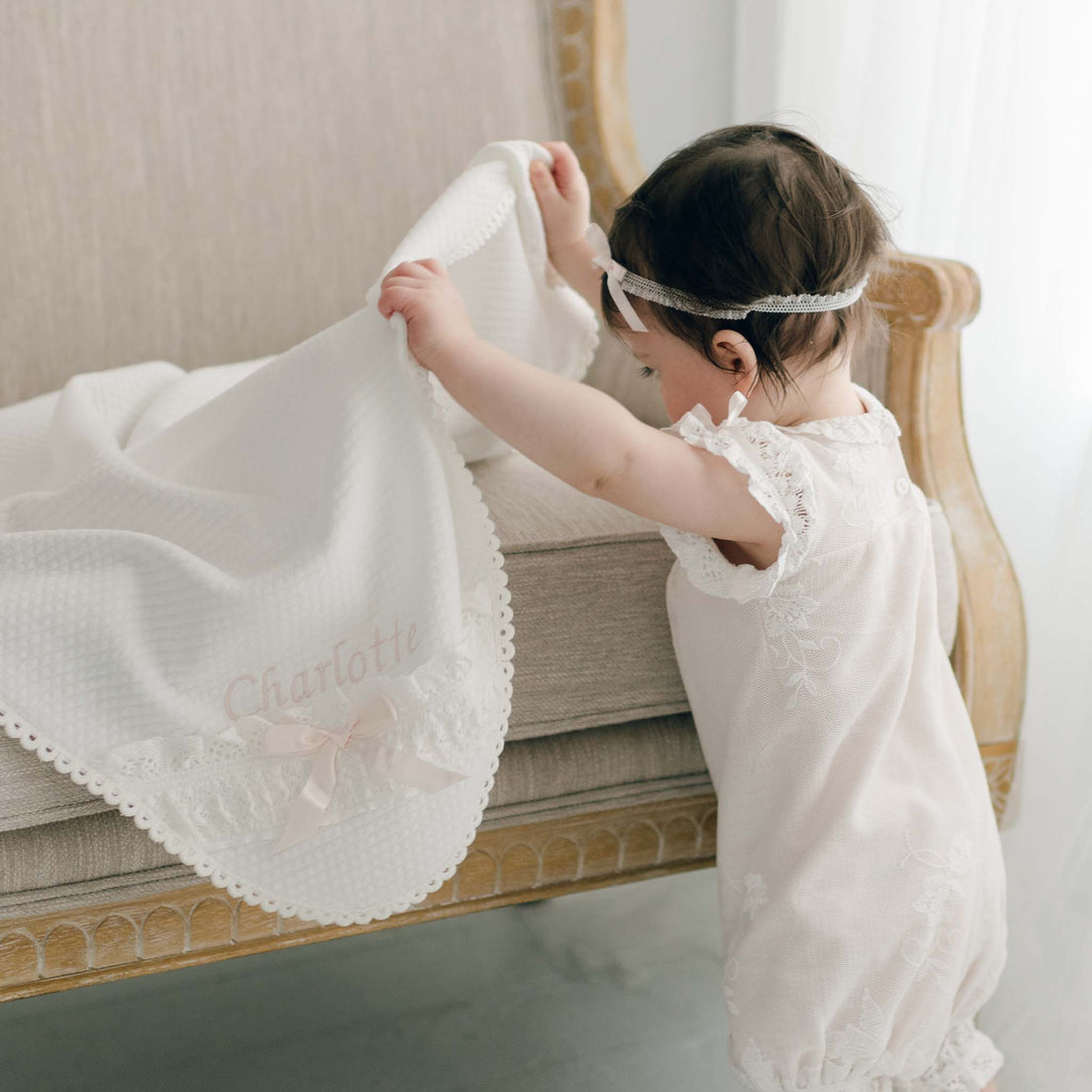 A baby girl in a Charlotte Romper and Lace Headband stands next to a beige couch, playfully lifting the Charlotte Personalized Blanket.