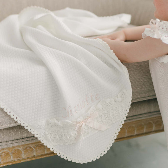 A baby's holds onto a beige upholstered chair that is draped with the Charlotte Personalized Blanket. The blanket features lace and ribbon details with the personalized name embroidery "Charlotte".