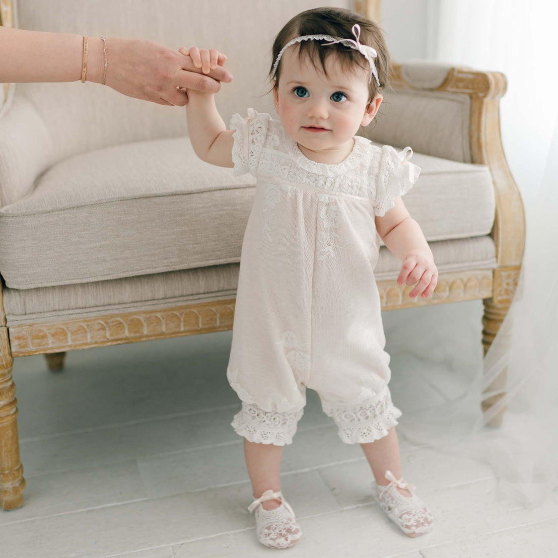 A baby girl with dark hair, wearing the Charlotte Romper with embroidered cotton lace and matching shoes, is holding an adult's hand while standing. The child has a headband with a small bow and is standing in front of a beige, upholstered chair with wooden accents.