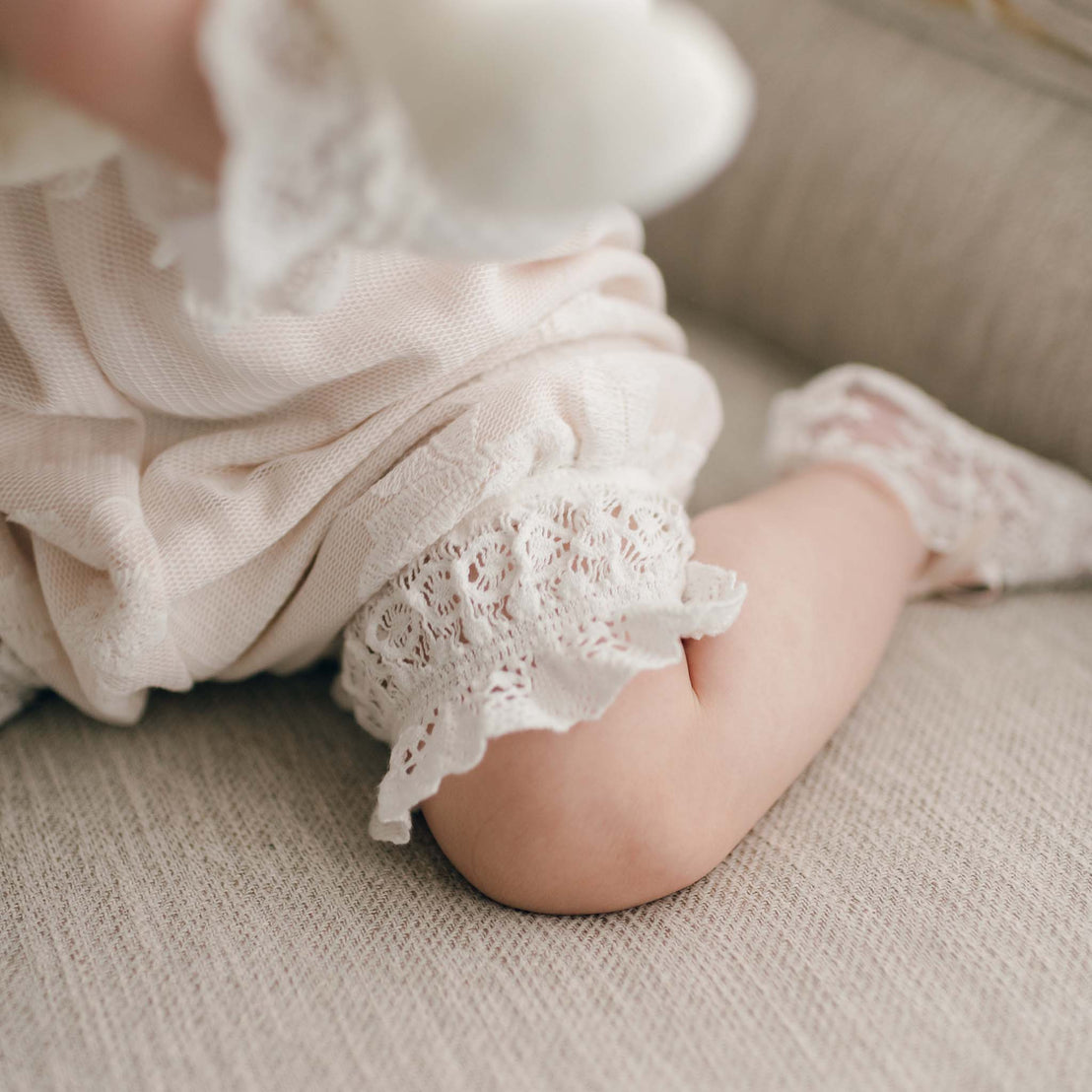 Close-up of a baby girl wearing a lace-trimmed Charlotte Romper, sitting on a beige fabric surface. The delicate outfit features intricate embroidered cotton lace designs.