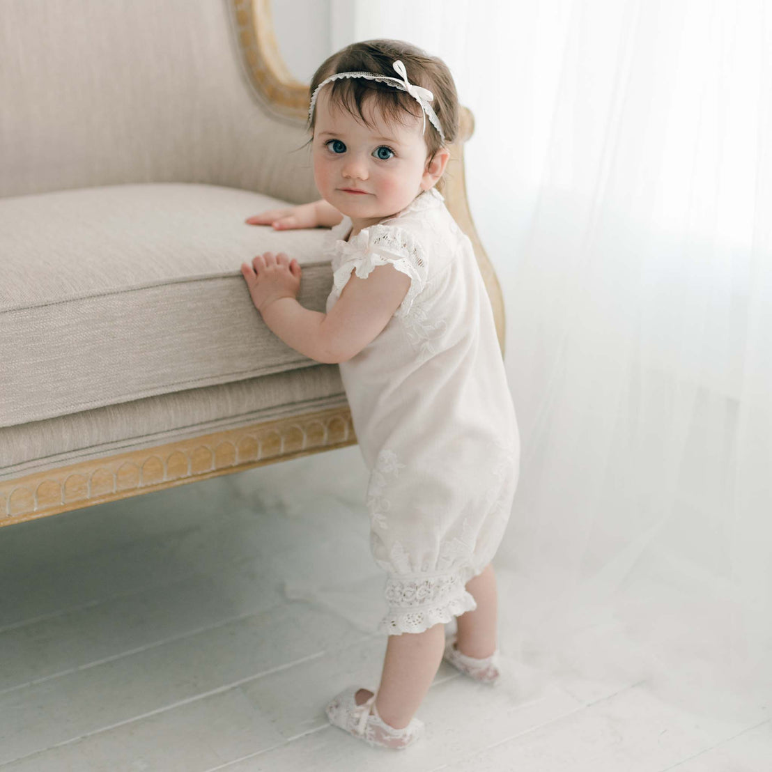 A baby girl with short brown hair, wearing a Charlotte Romper and a lace headband with a bow, stands while holding onto the seat of a cream-colored armchair. The background is softly lit with white curtains.